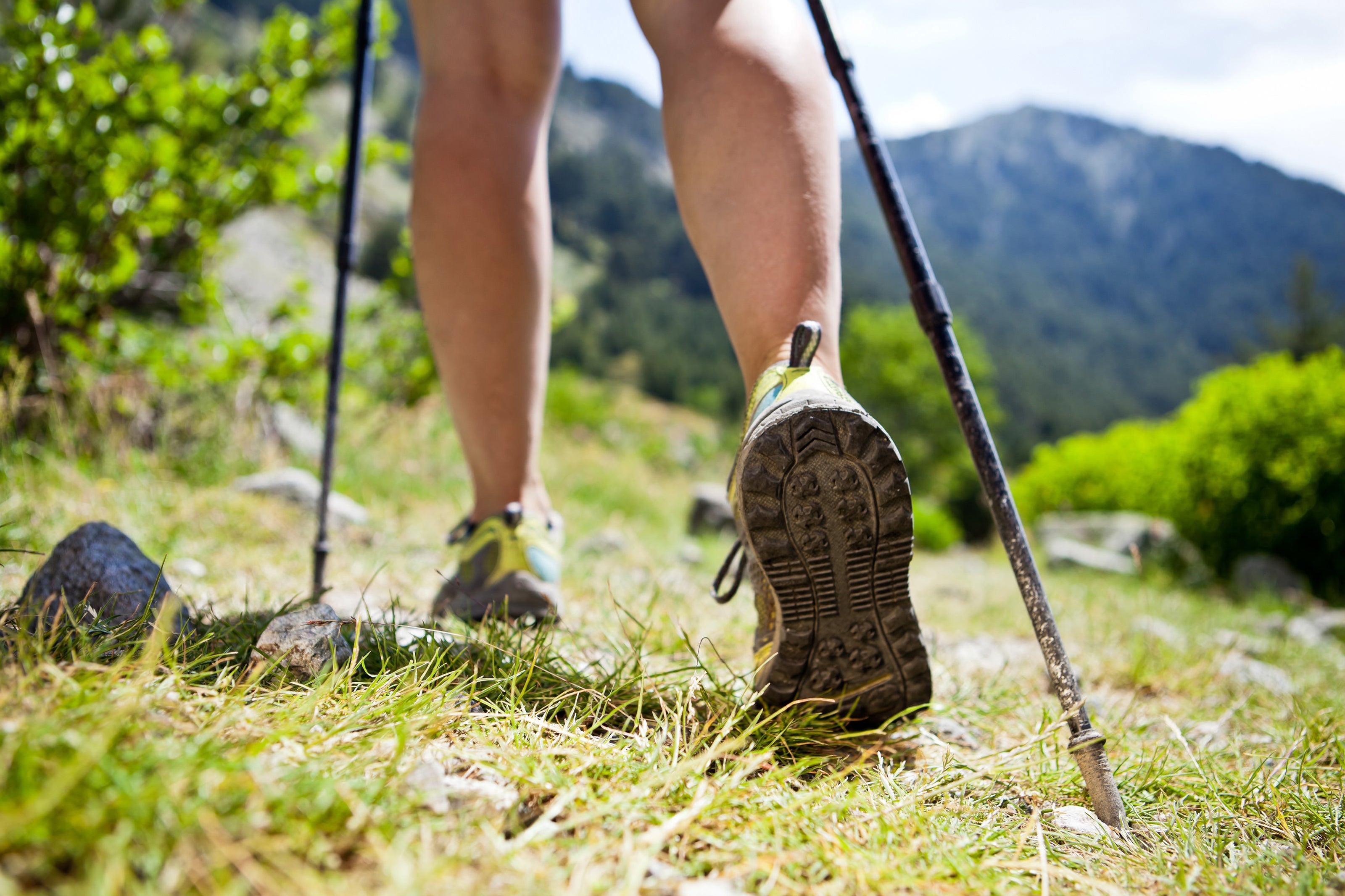 Mann in kurzen Hosen und Wanderstöcken läuft in Sportschuhe über eine karge Wiese